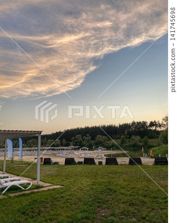 Beach area with sun loungers and umbrellas near a lake under a dramatic evening sky Beach area with sun loungers and umbrellas near a lake under a dramatic evening sky 141745698