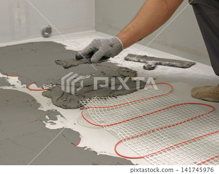 Worker installing electric underfloor heating system mat. Underfloor Heating Installation. Builder applying concrete mortar adhesive with trowel on cables. Energy Efficiency, Smart Home Foundation. 141745976