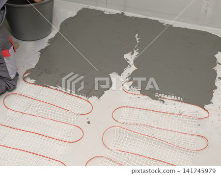 Worker installing electric underfloor heating system mat. Underfloor Heating Installation. Builder applying concrete mortar adhesive with trowel on cables. Energy Efficiency, Smart Home Foundation. Worker installing electric underfloor heating system mat. Underfloor Heating Installation. Builder applying concrete mortar adhesive with trowel on cables. Energy Efficiency, Smart Home Foundation. 141745979