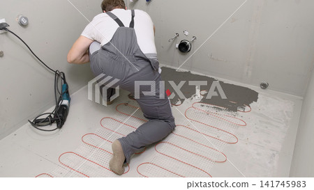 Worker installing electric underfloor heating system mat. Underfloor Heating Installation. Builder applying concrete mortar adhesive with trowel on cables. Energy Efficiency, Smart Home Foundation. Worker installing electric underfloor heating system mat. Underfloor Heating Installation. Builder applying concrete mortar adhesive with trowel on cables. Energy Efficiency, Smart Home Foundation. 141745983