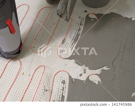 Worker installing electric underfloor heating system mat. Underfloor Heating Installation. Builder applying concrete mortar adhesive with trowel on cables. Energy Efficiency, Smart Home Foundation. Worker installing electric underfloor heating system mat. Underfloor Heating Installation. Builder applying concrete mortar adhesive with trowel on cables. Energy Efficiency, Smart Home Foundation. 141745986