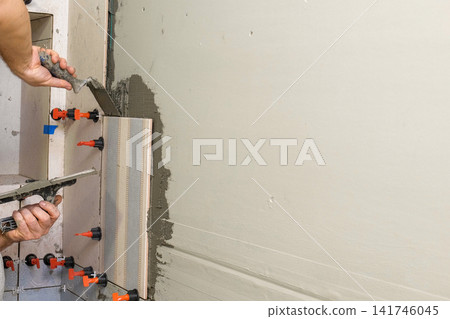 Professional tiler installing ceramic wall tiles with adhesive mortar. Worker hands using leveling system clips. Tiler hand adjusting tile. Wall tiling process. Interior Renovation. Precision Building Professional tiler installing ceramic wall tiles with adhesive mortar. Worker hands using leveling system clips. Tiler hand adjusting tile. Wall tiling process. Interior Renovation. Precision Building 141746045