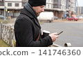 Bearded adult man using smartphone outdoors city street. Focused male looking at mobile phone screen, city road background, winter autumn commute. Urban Mobile Tech Digital Lifestyle. Remote Business 141746360