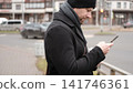 Bearded adult man using smartphone outdoors city street. Focused male looking at mobile phone screen, city road background, winter autumn commute. Urban Mobile Tech Digital Lifestyle. Remote Business 141746361