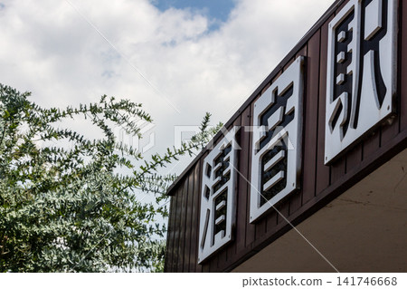 A view looking up at the station name sign and station building of Kiyone Station, against a bright blue sky and lush green scenery. A view looking up at the station name sign and station building of Kiyone Station, against a bright blue sky and lush green scenery. 141746668