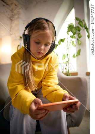 Focused school-age girl wearing headphones and gaming on smartphone Focused school-age girl wearing headphones and gaming on smartphone 141747727