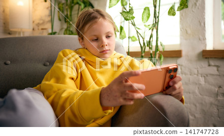 Preteen girl playing mobile game on smartphone while relaxing in armchair Preteen girl playing mobile game on smartphone while relaxing in armchair 141747728
