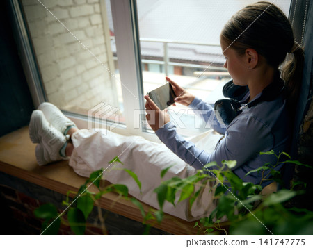 School-age girl browsing smartphone while resting beside window in modern apartment School-age girl browsing smartphone while resting beside window in modern apartment 141747775