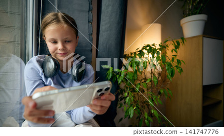 Happy school-age girl playing mobile game on smartphone in casual home setting Happy school-age girl playing mobile game on smartphone in casual home setting 141747776