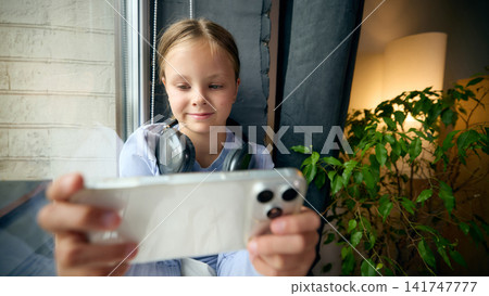 Focused preteen girl gaming on smartphone while wearing headphones at home Focused preteen girl gaming on smartphone while wearing headphones at home 141747777