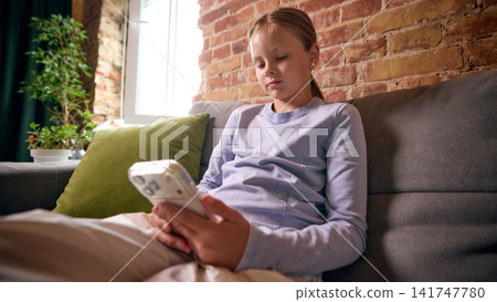 School-age girl scrolling smartphone while relaxing on sofa at home School-age girl scrolling smartphone while relaxing on sofa at home 141747780