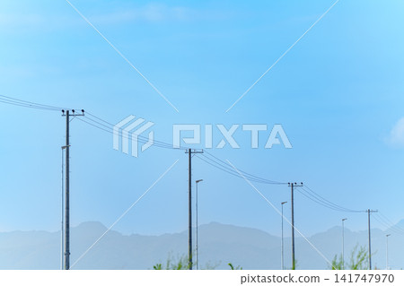 A nostalgic scene of utility poles and power lines stretching against a backdrop of blue sky and distant mountain ranges. A nostalgic scene of utility poles and power lines stretching against a backdrop of blue sky and distant mountain ranges. 141747970