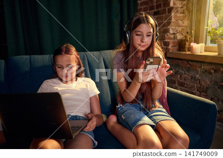 Two school-age girls using laptop and smartphone together on sofa Two school-age girls using laptop and smartphone together on sofa 141749009