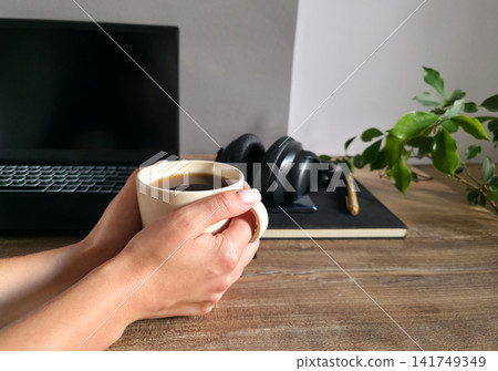 Hands holding a cup of coffee with a laptop, folded headphones and notebook blurred in the background. Concept of a work break and end of workday. Hands holding a cup of coffee with a laptop, folded headphones and notebook blurred in the background. Concept of a work break and end of workday. 141749349