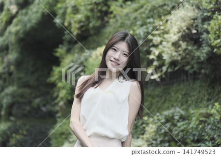 A young woman in a sleeveless dress smiles in a lush green park. 141749523
