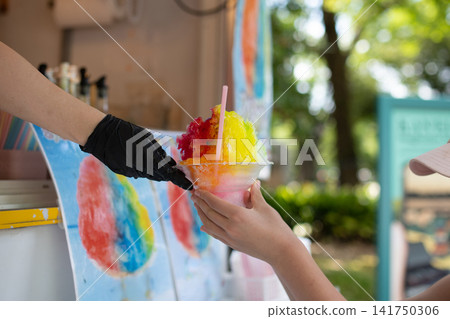 A child is handed a colorful rainbow shaved ice at a food stall. A child is handed a colorful rainbow shaved ice at a food stall. 141750306