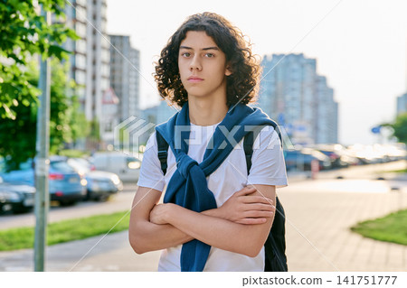 Serious teenage male student with curly hair and backpack standing on city street Serious teenage male student with curly hair and backpack standing on city street 141751777