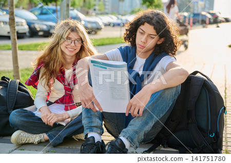Two teenage students sitting outdoors reading workbook preparing for exams together 141751780