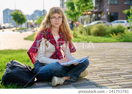 Thoughtful blonde student girl with glasses sitting on sidewalk holding notebook Thoughtful blonde student girl with glasses sitting on sidewalk holding notebook 141751781
