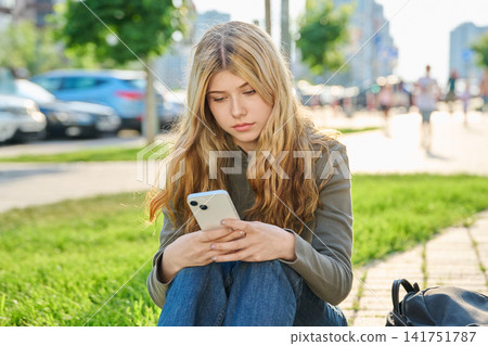 Serious teenage girl with long blonde hair sitting outdoors using smartphone Serious teenage girl with long blonde hair sitting outdoors using smartphone 141751787