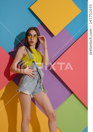 Young woman in yellow top and denim overalls posing against colorful geometric wall Young woman in yellow top and denim overalls posing against colorful geometric wall 141752035