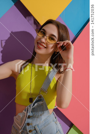 Young woman in yellow top and denim overalls smiling against colorful geometric wall 141752036