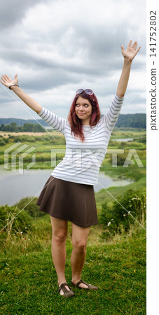 Young woman poses with hands raised on a hillside near a river under cloudy sky Young woman poses with hands raised on a hillside near a river under cloudy sky 141752802