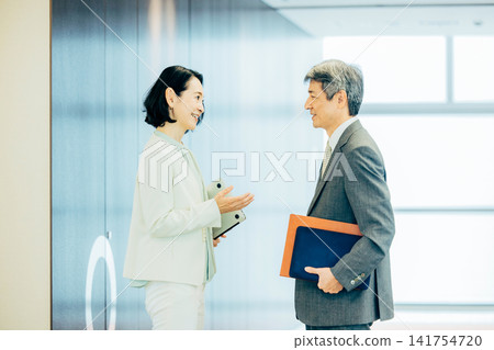 身著西裝的高級男女在辦公室走廊交談。 身著西裝的高級男女在辦公室走廊交談。 141754720