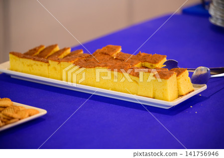 Sliced sponge cake served on a white plate during a catered event buffet on a blue tablecloth with metal tongs. Sliced sponge cake served on a white plate during a catered event buffet on a blue tablecloth with metal tongs. 141756946