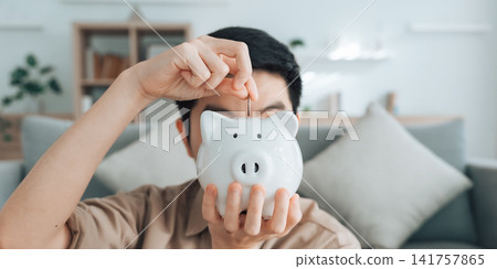Hand of young Asian man putting coin in piggy bank. Save money and financial investment Hand of young Asian man putting coin in piggy bank. Save money and financial investment 141757865