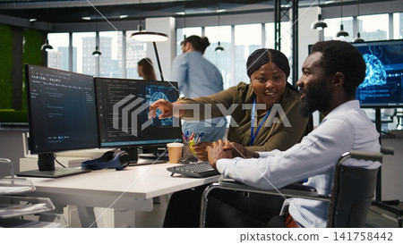 Cheerful manager and employee with paraplegia in inclusive workplace developing software applications. African american technician and colleague with disability using AI to solve bottlenecks, camera A Cheerful manager and employee with paraplegia in inclusive workplace developing software applications. African american technician and colleague with disability using AI to solve bottlenecks, camera A 141758442