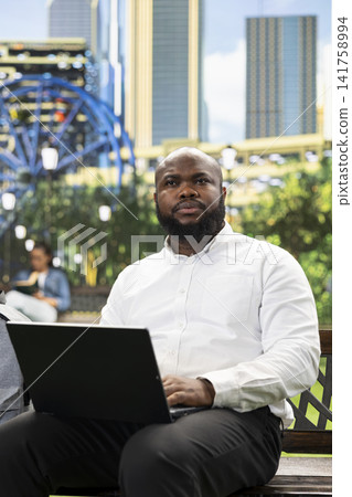 Businessman looking thoughtful beyond laptop while seated on city park bench, capturing moment of planning during flexible outdoor work. Pensive professional with remote job. Businessman looking thoughtful beyond laptop while seated on city park bench, capturing moment of planning during flexible outdoor work. Pensive professional with remote job. 141758994