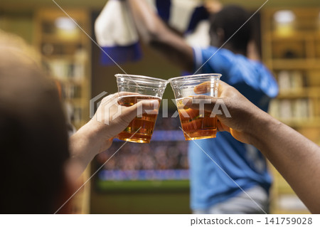 Close up of football supporters drinking beers together while watching match on TV. Back view of fans enjoying alcohol drinks in living room during live game broadcast Close up of football supporters drinking beers together while watching match on TV. Back view of fans enjoying alcohol drinks in living room during live game broadcast 141759028