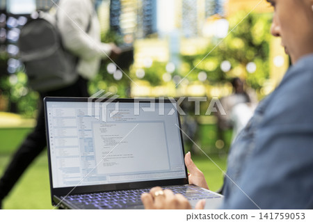Close up of person working with programming interface and source code on laptop, representing software development and flexible IT work in city park. Over-the-shoulder view. Close up of person working with programming interface and source code on laptop, representing software development and flexible IT work in city park. Over-the-shoulder view. 141759053