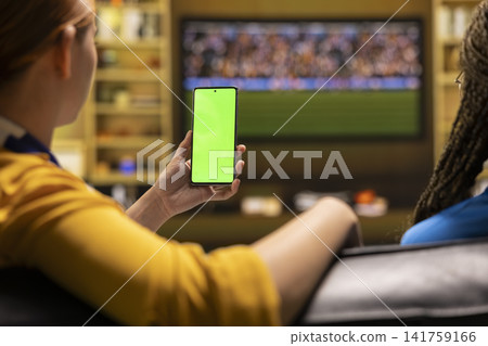 Green screen phone used by football fan watching match on TV at home. Soccer supporter holding mockup smartphone in living room watching live game, close up Green screen phone used by football fan watching match on TV at home. Soccer supporter holding mockup smartphone in living room watching live game, close up 141759166