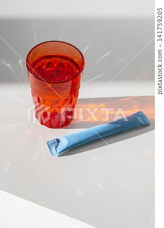 Red glass of water with light reflections and blue sachet on white surface. Red glass of water with light reflections and blue sachet on white surface. 141759205