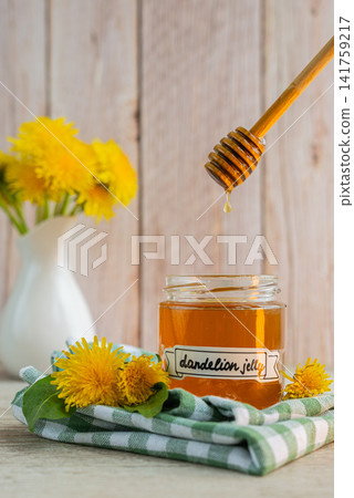 Transparent glass of jar dandelion jelly wooden honey dipper coated with jelly dripping with dandelion flowers in vase. Soft natural lighting highlights the amber color glossy texture and artisanal Transparent glass of jar dandelion jelly wooden honey dipper coated with jelly dripping with dandelion flowers in vase. Soft natural lighting highlights the amber color glossy texture and artisanal 141759217
