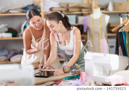 Two female dressmakers drawing a model of clothes on a tablet Two female dressmakers drawing a model of clothes on a tablet 141759323
