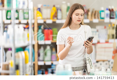Female shopper searches for necessary goods on the shelves of supermarket, checking the list on her smartphone 141759413