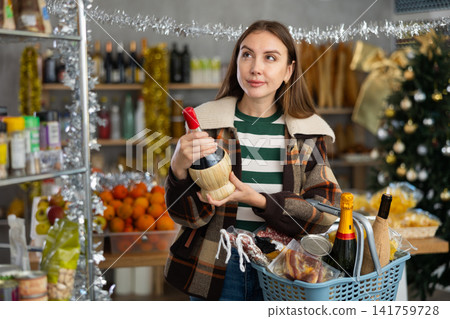 Female shopper with grocery cart chooses wine for celebrating Christmas or New Year Female shopper with grocery cart chooses wine for celebrating Christmas or New Year 141759728