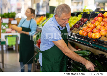 Focused mature salesman laying out small green cucumbers in supermarket Focused mature salesman laying out small green cucumbers in supermarket 141759786