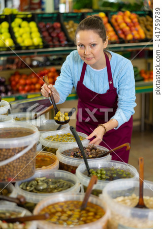 Positive middle-aged female merchandiser in apron picking up olives with scoop from plastic container in grocery store Positive middle-aged female merchandiser in apron picking up olives with scoop from plastic container in grocery store 141759789