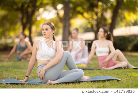 Sporty young girls performing seated twisting asana Matsyendrasana on mat in in summer park during daily yoga session Sporty young girls performing seated twisting asana Matsyendrasana on mat in in summer park during daily yoga session 141759826