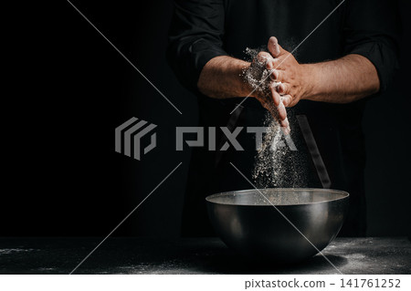Baker dusting flour from hands above mixing bowl on dark background Baker dusting flour from hands above mixing bowl on dark background 141761252
