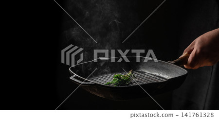 Chef holding hot cast iron grill pan with rosemary and steam on dark background Chef holding hot cast iron grill pan with rosemary and steam on dark background 141761328