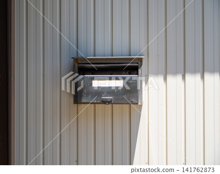 A metal mailbox mounted on a bright corrugated sheet metal exterior wall. Sunlight casts shadows on the uneven surface of the wall, creating a simple yet inviting look. A metal mailbox mounted on a bright corrugated sheet metal exterior wall. Sunlight casts shadows on the uneven surface of the wall, creating a simple yet inviting look. 141762873