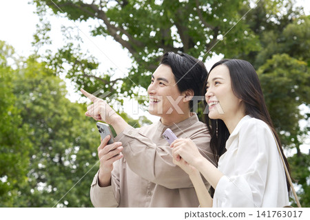 A smiling couple in their 30s talking on their smartphones in a park. 141763017