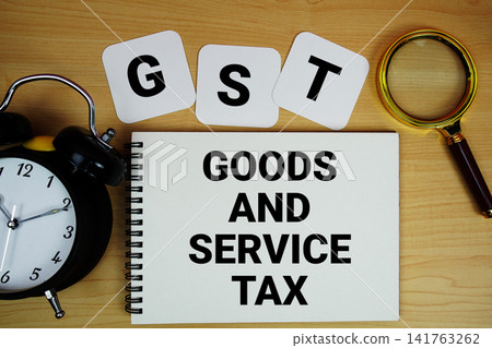 GST Goods and Service Tax text written on notepad with magnifying glass and alarm clock on wooden background GST Goods and Service Tax text written on notepad with magnifying glass and alarm clock on wooden background 141763262