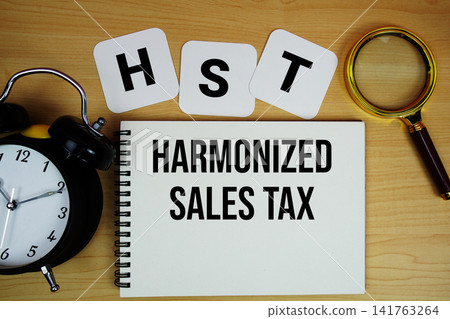 HST Harmonized Sales Tax text written on notepad with magnifying glass and alarm clock on wooden background 141763264