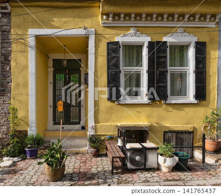 Sunny Mediterranean yellow house facade with a traditional door, black shutters, decorative trim, and potted plants Sunny Mediterranean yellow house facade with a traditional door, black shutters, decorative trim, and potted plants 141765437